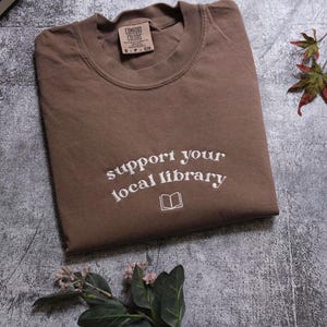 a brown t-shirt with the text "Support your local library" printed on it, along with a small book icon. The t-shirt is placed on a gray surface, and there is a small plant and a leaf in the foreground.