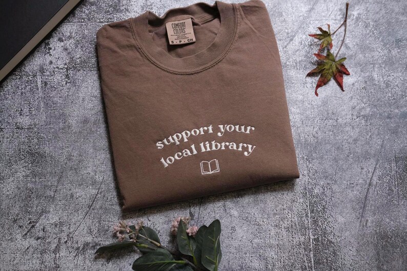 a brown t-shirt with the text "Support your local library" printed on it, along with a small book icon. The t-shirt is placed on a gray surface, and there is a small plant and a leaf in the foreground.