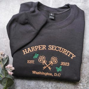 A black sweatshirt with the text "HARPER SECURITY EST. 2023 Washington, D.C." printed on it, along with a key design and two butterflies.