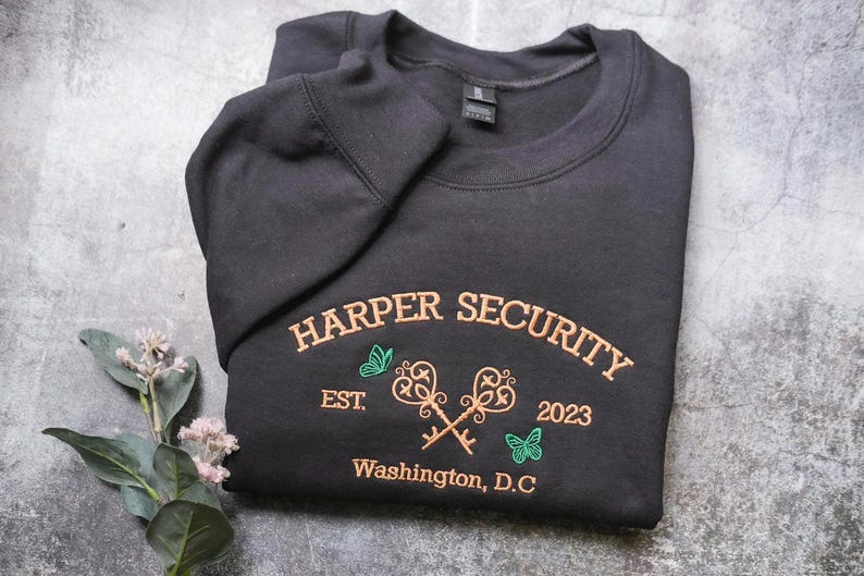 A black sweatshirt with the text "HARPER SECURITY EST. 2023 Washington, D.C." printed on it, along with a key design and two butterflies.