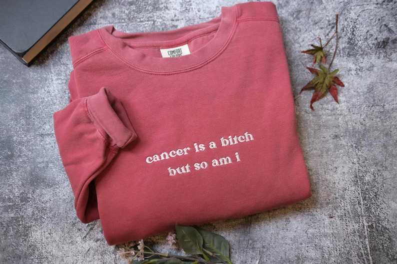 A red long-sleeved shirt with the text "cancer is a shit but so am I" printed on it, along with some leaves and a book.