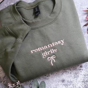 a green sweatshirt with the text "romantasty girlie" embroidered on it, along with a bow detail. The sweatshirt is folded and placed on a gray surface, with some leaves and a plant visible in the background.