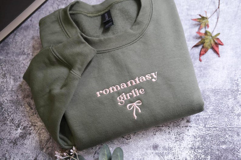 a green sweatshirt with the text "romantasty girlie" embroidered on it, along with a bow detail. The sweatshirt is folded and placed on a gray surface, with some leaves and a plant visible in the background.