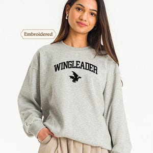 A young woman wearing a grey sweatshirt with the text "WINGLEADER" printed on it, standing against a white background.