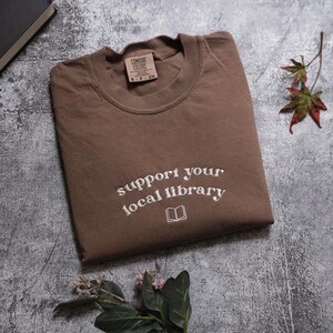 a brown t-shirt with the text "Support your local library" printed on it, along with a leaf and a book.