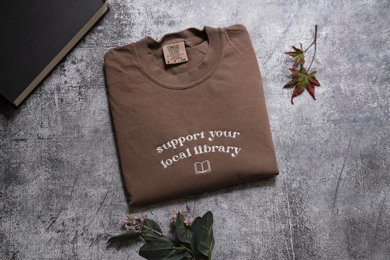 a brown t-shirt with the text "Support your local library" printed on it, along with a leaf and a book.