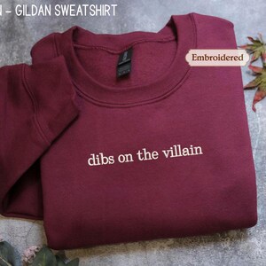 A maroon-colored GILARD sweatshirt with the text "dibs on the villain" embroidered on it, along with a leaf and a tag.