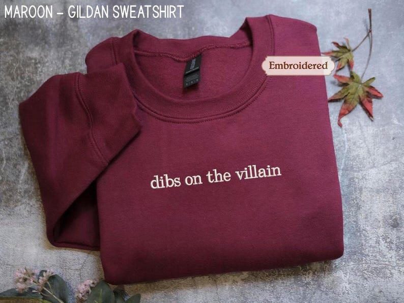 A maroon-colored GILARD sweatshirt with the text "dibs on the villain" embroidered on it, along with a leaf and a tag.