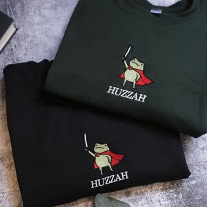 May include: Two sweatshirts, one black and one dark green, are displayed. Each features an embroidered cartoon frog wearing a red cape and holding a sword. The word "Huzzah" is embroidered below the frog. The sweatshirts are on a textured surface.