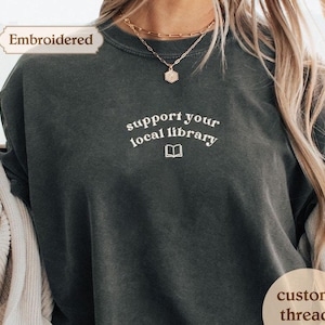 A person wearing a gray t-shirt with the text "Support your local library" printed on it, along with a necklace and a sweater.