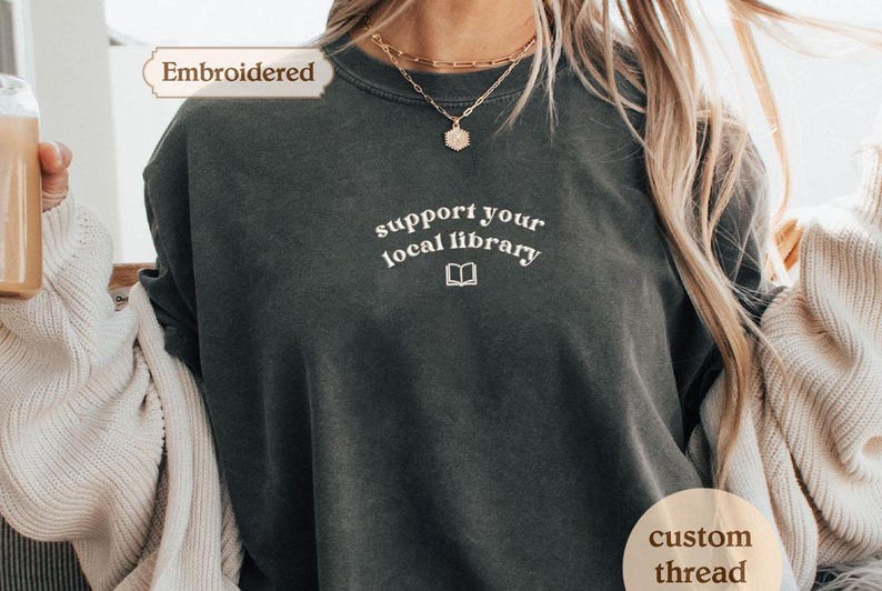 A person wearing a gray t-shirt with the text "Support your local library" printed on it, along with a necklace and a sweater.