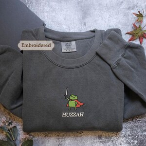 a gray sweatshirt with a green frog character and the word "HUZZAH" printed on it, along with a label that reads "Embroidered".
