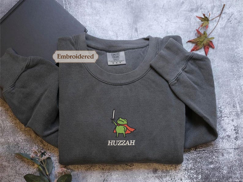 a gray sweatshirt with a green frog character and the word "HUZZAH" printed on it, along with a label that reads "Embroidered".