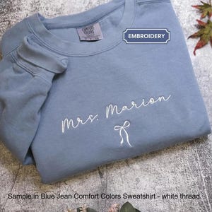May include: A blue jean coloured crewneck sweatshirt featuring white embroidered text that reads Mrs. Marion above a small white embroidered bow graphic. The garment includes a neck label and a rectangular tag labelled EMBROIDERY.