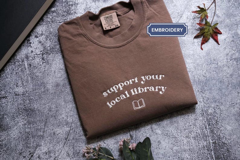 A brown t-shirt with the text "Support your local library" printed on it, along with a small book icon, is folded and placed on a gray surface. There are some leaves and flowers in the background.