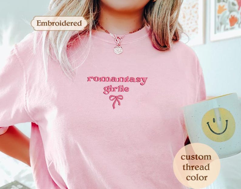 A person wearing a pink t-shirt with the text "romancey girl" embroidered on it, holding a mug with a smiley face on it.