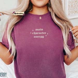 A person with long blonde hair wearing a purple t-shirt with the text "main character energy" embroidered on it.