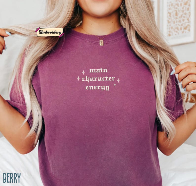 A person with long blonde hair wearing a purple t-shirt with the text "main character energy" embroidered on it.