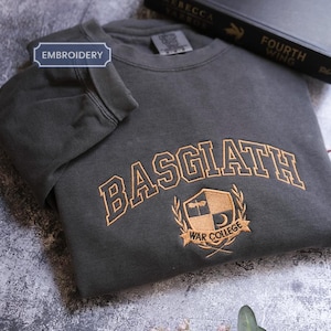 a gray sweatshirt with the word "BASGLATH" embroidered on it, along with a black book titled "FOURTH WING" and a leaf on the surface.