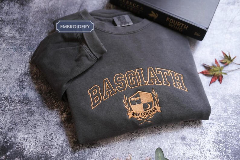 a gray sweatshirt with the word "BASGLATH" embroidered on it, along with a black book titled "FOURTH WING" and a leaf on the surface.