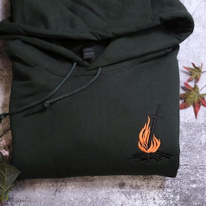 a black hoodie with an embroidered design depicting a campfire on the front.
