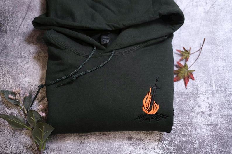 a black hoodie with an embroidered design depicting a campfire on the front.