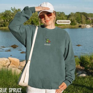 A woman wearing a green sweatshirt with a frog design and sunglasses stands in front of a body of water, with a boat visible in the background.