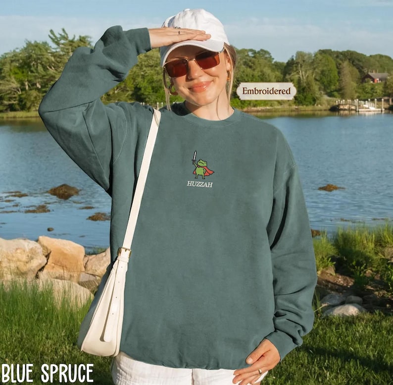 A woman wearing a green sweatshirt with a frog design and sunglasses stands in front of a body of water, with a boat visible in the background.