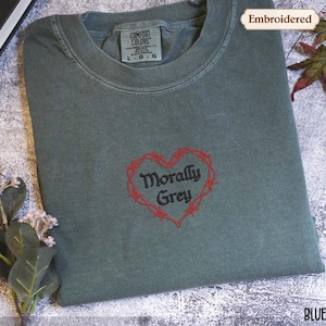 a gray t-shirt with a red heart-shaped embroidered design and the text "Morally Grey" printed on it. The t-shirt is placed on a white surface, and there are some leaves and flowers nearby.