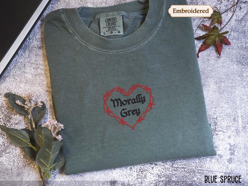 a gray t-shirt with a red heart-shaped embroidered design and the text "Morally Grey" printed on it. The t-shirt is placed on a white surface, and there are some leaves and flowers nearby.