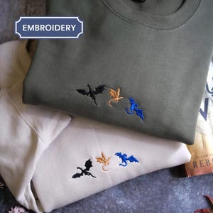 May include: Two sweatshirts, one olive green and one cream, each embroidered with three small dragon silhouettes in black, gold, and blue. The word "EMBROIDERY" is in a blue box. A book titled "Fourth Wing" is visible.