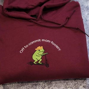 May include: A maroon hooded sweatshirt featuring an embroidered graphic of a green frog wearing a yellow hat and riding a black scooter. The text "Off to commit mom foolery" is embroidered in white above the frog illustration.