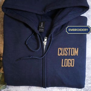 Peut inclure: Sweat à capuche bleu marine avec fermeture éclair argentée. Le texte "CUSTOM LOGO" est brodé en or sur le devant. Le mot "EMBROIDERY" est dans une étiquette bleue et dorée.