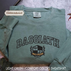 A light green, embroidered sweatshirt with the text "BASGIATH WARREN COLLEGE" printed on it, along with a label indicating it is an "Embroidered" item. The sweatshirt is placed on a gray surface, with a leaf and a plant visible in the background.