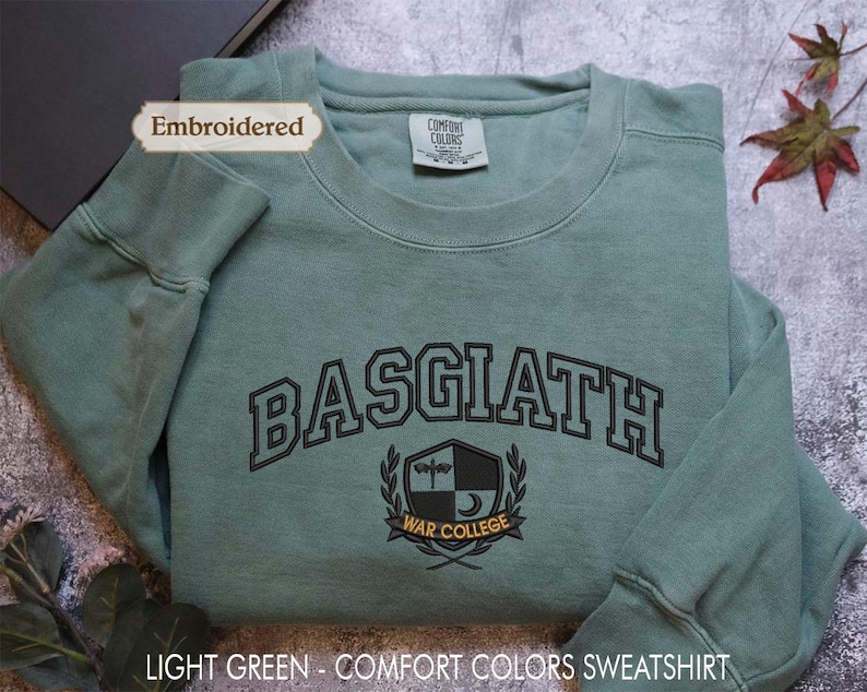 A light green, embroidered sweatshirt with the text "BASGIATH WARREN COLLEGE" printed on it, along with a label indicating it is an "Embroidered" item. The sweatshirt is placed on a gray surface, with a leaf and a plant visible in the background.
