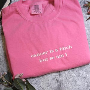 A pink t-shirt with the text "cancer is a bitch but so am I" printed on it, along with some leaves and a book.