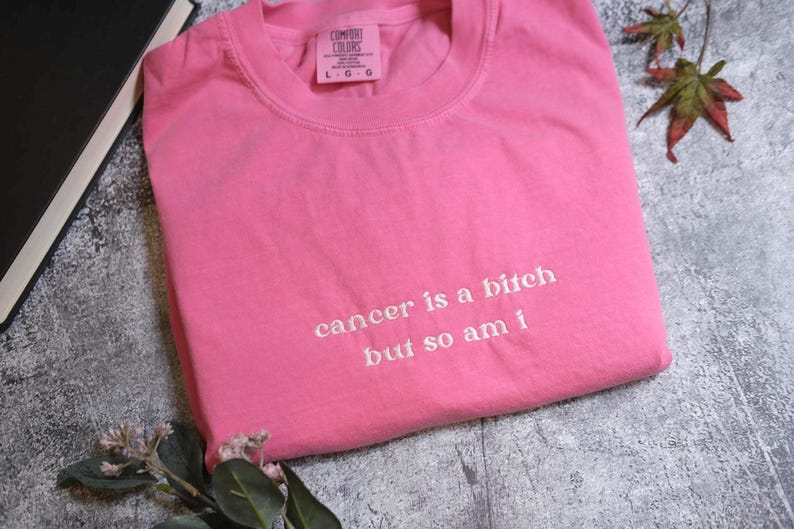 A pink t-shirt with the text "cancer is a bitch but so am I" printed on it, along with some leaves and a book.