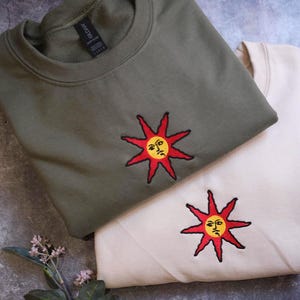 two t-shirts with sun-like embroidered designs, one in a dark green color and the other in a lighter beige color, placed on a gray surface alongside some leaves.