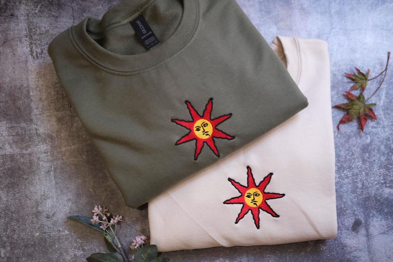 two t-shirts with sun-like embroidered designs, one in a dark green color and the other in a lighter beige color, placed on a gray surface alongside some leaves.