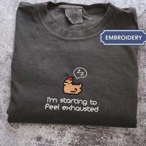May include: Dark grey t-shirt featuring an embroidered pixelated chicken design. The chicken has a red comb and is sleeping, with a speech bubble above it saying "zz". The text "I'm starting to feel exhausted" is below the chicken.