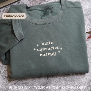 a gray sweatshirt with the text "main character energy" embroidered on it. The sweatshirt is folded and placed on a gray surface, with a few leaves visible in the background.
