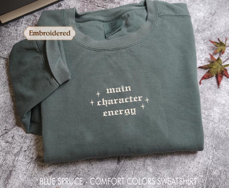 a gray sweatshirt with the text "main character energy" embroidered on it. The sweatshirt is folded and placed on a gray surface, with a few leaves visible in the background.