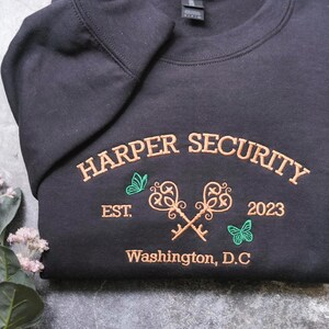 A black t-shirt with gold text and graphics, including the words "Harper Security" and "Est. 2023 Washington, D.C."