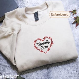 A white t-shirt with a heart-shaped embroidered design and the text "Morally Grey" on it, along with a tag that says "Embroidered". The t-shirt is placed on a gray surface, and there is a book and some leaves in the background.