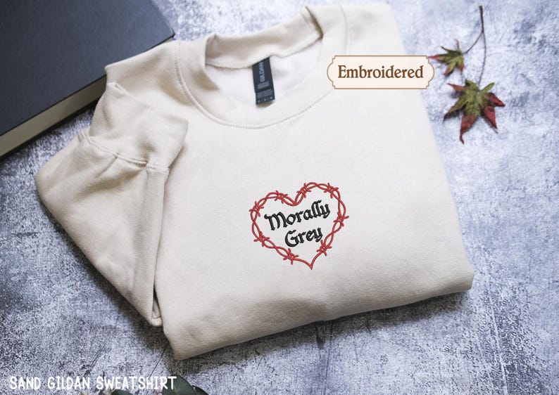 A white t-shirt with a heart-shaped embroidered design and the text "Morally Grey" on it, along with a tag that says "Embroidered". The t-shirt is placed on a gray surface, and there is a book and some leaves in the background.