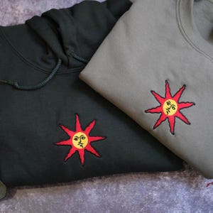 two sweatshirts with sun-like embroidered designs on them, one black and one gray.