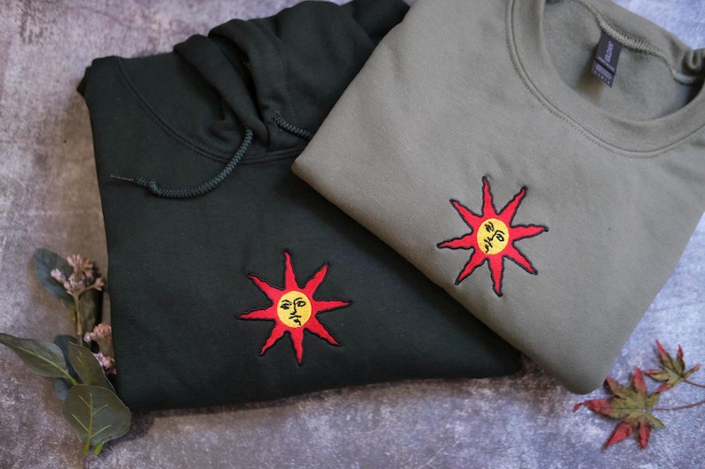 two sweatshirts with sun-like embroidered designs on them, one black and one gray.