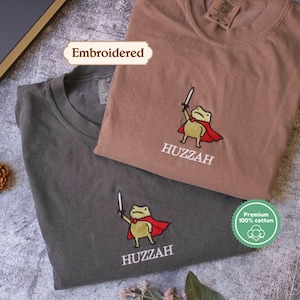 two t-shirts with a frog character, one in a brown shirt and the other in a gray shirt, both with the word "Huzzah" printed on them.