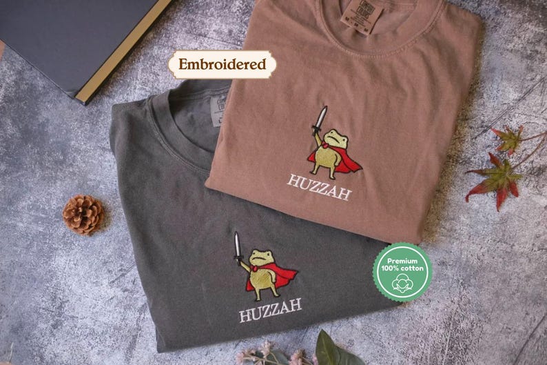 two t-shirts with a frog character, one in a brown shirt and the other in a gray shirt, both with the word "Huzzah" printed on them.