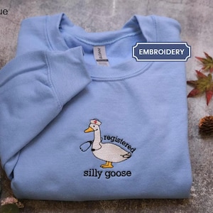 May include: A Carolina blue crewneck sweatshirt featuring an embroidered graphic of a white goose wearing a nurse cap and a stethoscope. The text "registered silly goose" is embroidered in black below the goose. A label reads "EMBROIDERY".